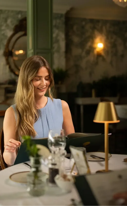 Elegant dining room with smiling woman in blue dress, ambient lighting, and green decor.