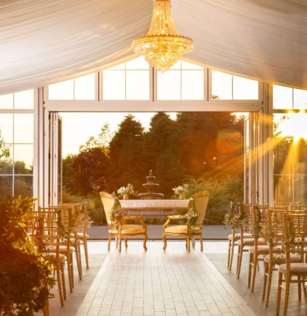 Elegant indoor wedding venue with sunlight streaming through large windows, greenery, and fountain.