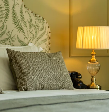 Elegant hotel room with cosy bed, fern-print headboard, and warm-lit lamp.