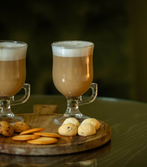 Two mugs of frothy coffee with assorted cookies on a wooden tray in a cosy setting.