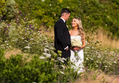 Couple in wedding attire smiling amidst lush garden blooms.