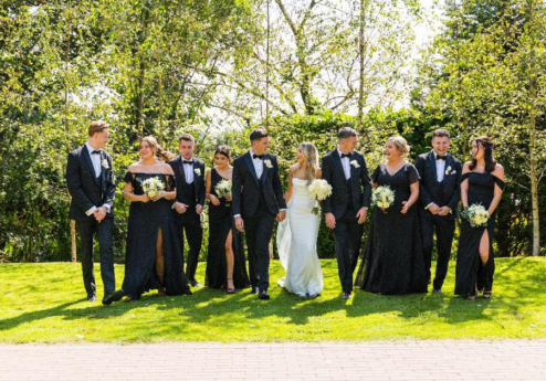 A wedding party poses joyfully outdoors in formal attire, surrounded by lush greenery.