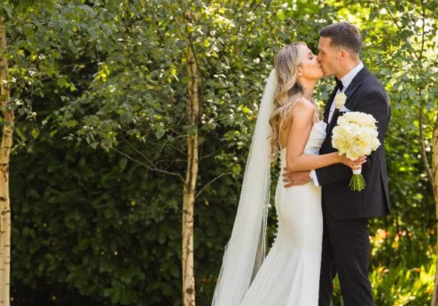 A couple sharing a kiss in a lush, green garden at Fernhill House Hotel, with the bride in a white dress holding a bouquet.