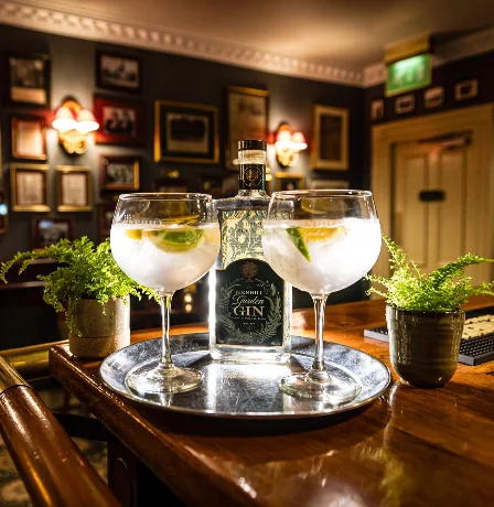 Cosy hotel bar with framed photos, glowing lamps, and two gin glasses on a tray.