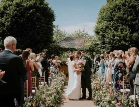 A couple kisses at an outdoor wedding ceremony surrounded by family and blooming flowers.