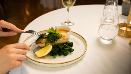 Elegant dinner setting with a person enjoying grilled fish, greens, and a glass of white wine.