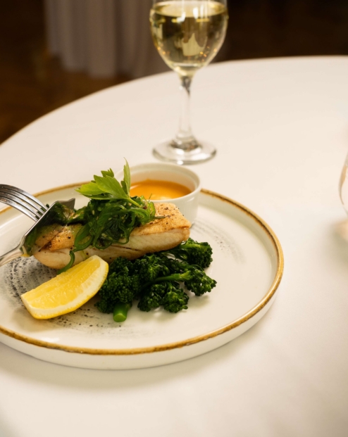 Elegant dinner setting with a person enjoying grilled fish, greens, and a glass of white wine.