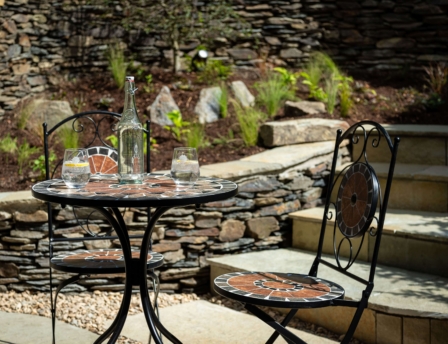 Cosy outdoor seating with a mosaic table, two glasses of water, and a stone wall backdrop.