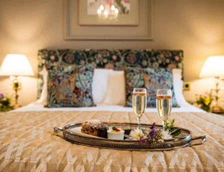 Cosy hotel room with a king bed, floral pillows, and a tray of desserts and champagne.