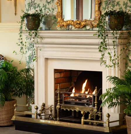 Cosy hotel lounge with a warm fireplace surrounded by lush greenery.