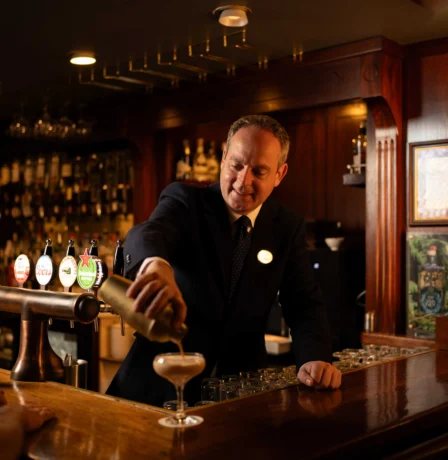 Bartender serving drinks in cosy Fernhill House Hotel bar, with a guest at the counter.