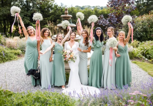 Bride and bridesmaids in sage dresses cheering by a fountain with bouquets, garden wedding