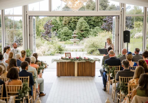 Family wedding ceremony indoors opening onto gardens with fountain, guests seated and celebrant speaking